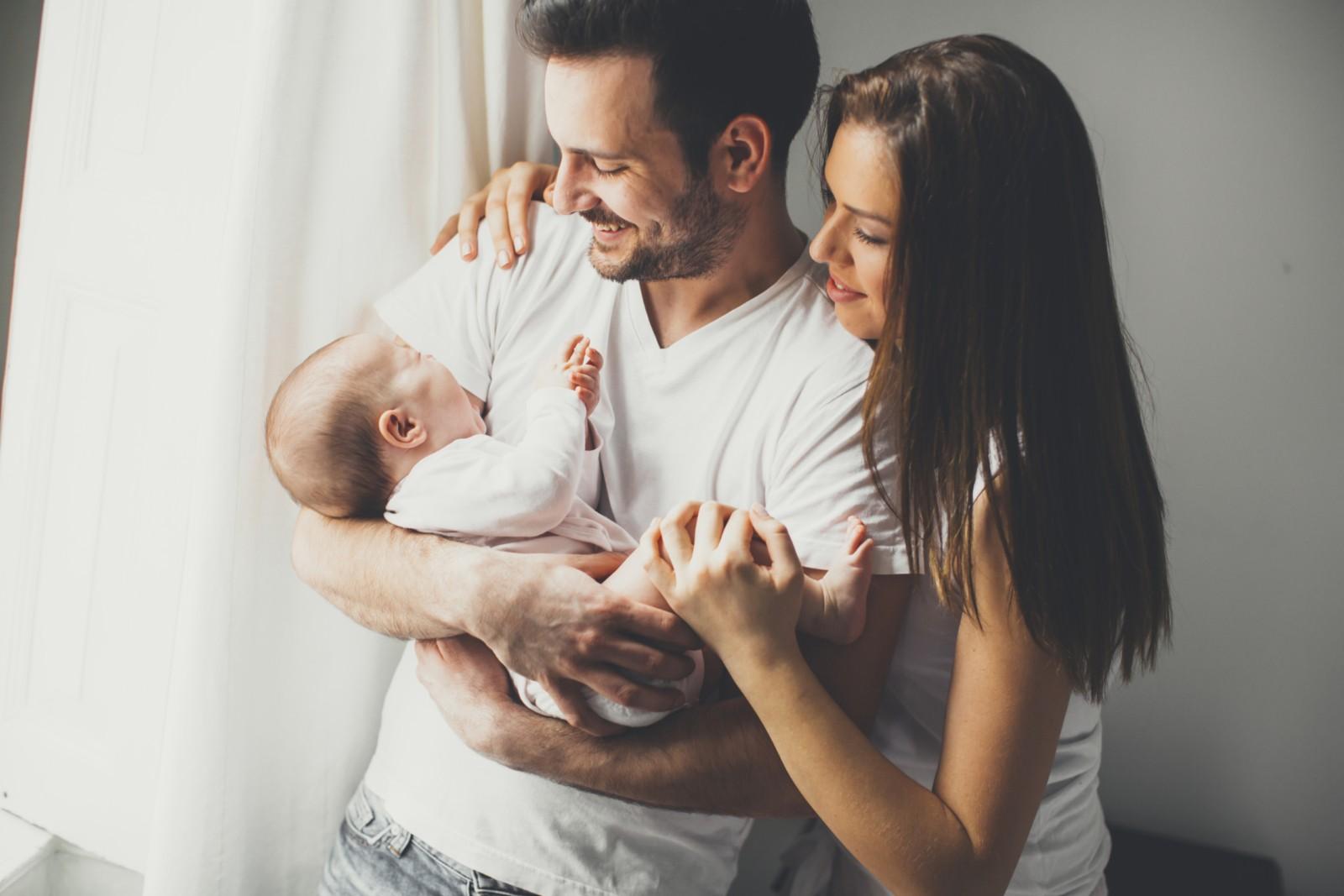 Ein junger Vater hält sein neu geborenes Baby auf dem Arm während er von seiner Partnerin umarmt wird © Shutterstock